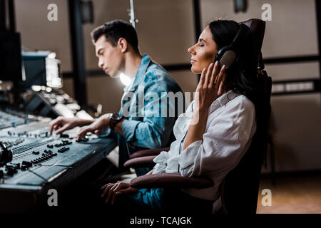 pretty sound producer in headphones listening music near mixed racial friend working at mixing console Stock Photo