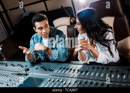 two smiling multicultural sound producers talking while sitting near mixing console in recording studio Stock Photo