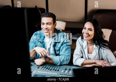 two cheerful multicultural sound producers working at mixing console in recording studio Stock Photo