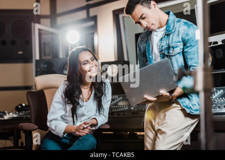 handsome mixed race sound producer using laptop near cheerful smiling colleague Stock Photo