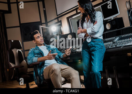 two young multicultural sound producers talking near mixing console in recording studio Stock Photo