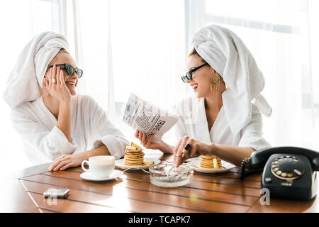 stylish happy women in bathrobes, sunglasses and jewelry with towels on heads smoking cigarette, laughing and reading newspaper at morning Stock Photo