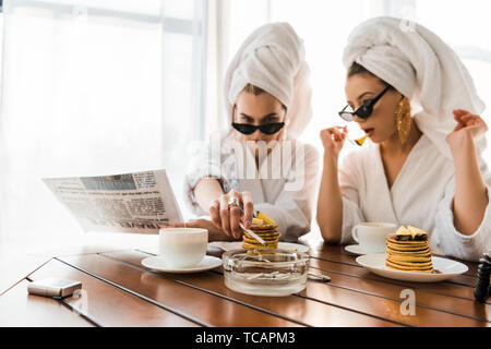 selective focus of stylish women in bathrobes, sunglasses and jewelry with towels on heads smoking cigarette and reading newspaper while eating pancak Stock Photo