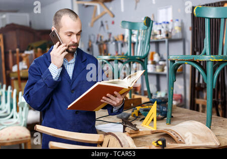 Professional furniture restorer holding album and talking on mobile phone in workshop Stock Photo