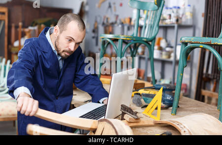 Professional young man carpenter using laptop while repairing antique furniture in workshop Stock Photo