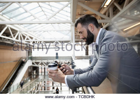 Businessman with coffee using smart phone on office atrium balcony