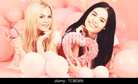 Valentines day concept. Girls lay near balloons, holds heart toys, pink background. Blonde and brunette on smiling faces dreaming about love and date. Sisters, friends in pajamas at pajamas party. Stock Photo