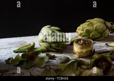 closeup of some whole raw fresh artichokes and some other cut artichokes and their leaves on a white rustic wooden table, against a black background Stock Photo