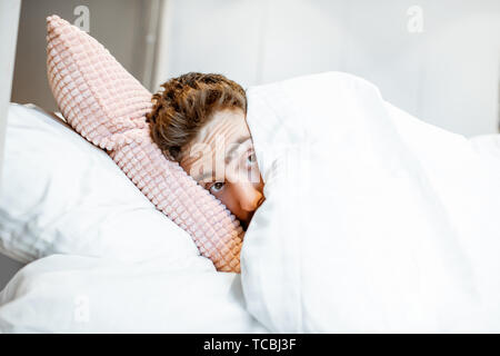 Man hiding in bed under the white sheets feeling scared alone Stock Photo