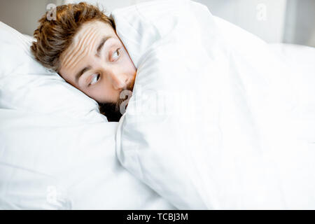 Man hiding in bed under the white sheets feeling scared alone Stock Photo