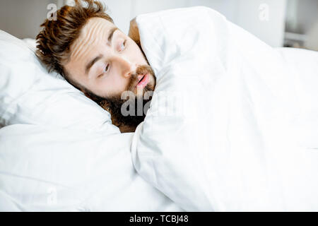 Man hiding in bed under the white sheets feeling scared alone Stock Photo