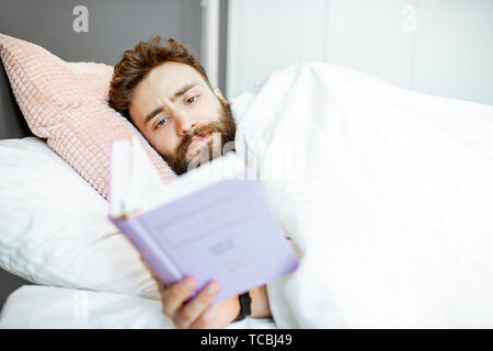 Creative man reading book before the sleep, lying under the white sheets at the bedroom Stock Photo