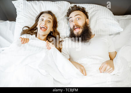 Portrait of a young couple feeling surprised and shocked lying together on the bed under the white sheets Stock Photo