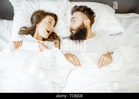 Portrait of a young couple feeling surprised and shocked lying together on the bed under the white sheets Stock Photo