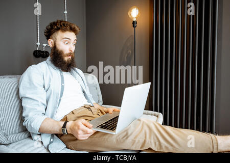 Creative bearded man with surprised emotions working with laptop while sitting on the couch in the living room at home Stock Photo