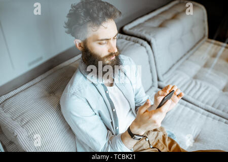 Creative bearded man using smart phone, relaxing on the couch at home Stock Photo
