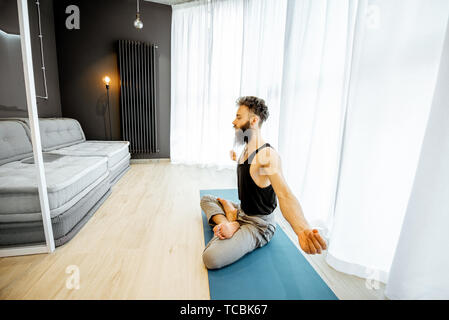 Young bearded man practising yoga, feeling relaxed, sitting in the lotus position in the living room at home Stock Photo