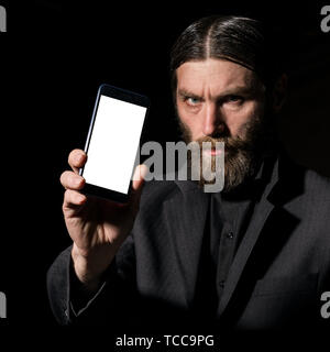 Old believer senior priest with a smartphone, bearded old man is calling on a dark background Stock Photo