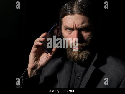 Old believer senior priest with a smartphone, bearded old man is calling on a dark background Stock Photo