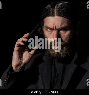 Old believer senior priest with a smartphone, bearded old man is calling on a dark background Stock Photo