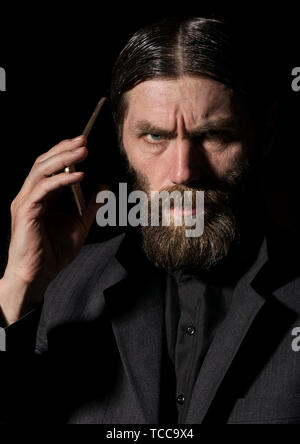 Old believer senior priest with a smartphone, bearded old man is calling on a dark background Stock Photo