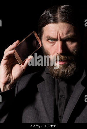 Old believer senior priest with a smartphone, bearded old man is calling on a dark background Stock Photo