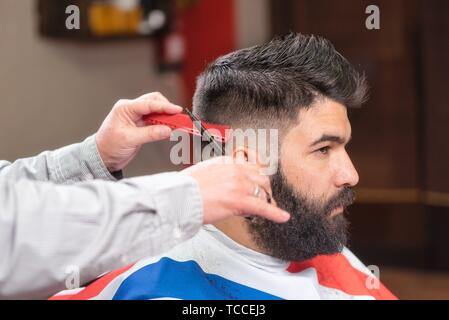 Handsome bearded man while having his hair cut by hairdresser at  barbershop. Close up side view of young bearded man getting groomed by  hairdresser with hair dryer at barbershop Stock Photo -