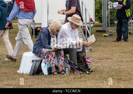 A retirement age couple sat on a bench reading during an outdoor concert one reading a book and one reading the newspaper Stock Photo