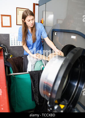 Glad woman in uniform taking out clothes from washing machine at laundry Stock Photo