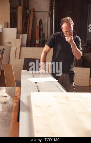 Carpenter or woodworker in his workshop standing looking thoughtfully down at the floor alongside his work bench with planks of wood Stock Photo