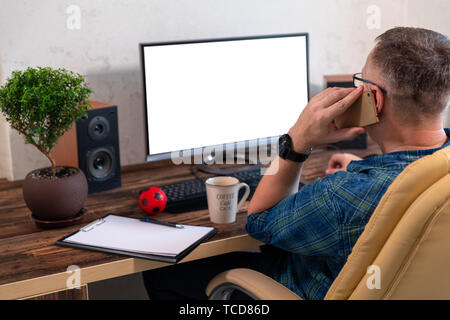 Businessman making a phone call at the office on his smartphone as he sits in front of a large blank desktop monitor with notepad alongside, rear side Stock Photo