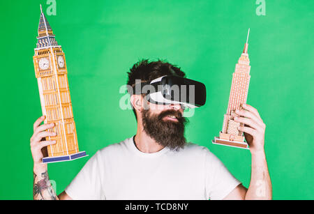 Man with beard in VR glasses, green background. 3D design concept. Guy  holds Big Ben and Empire State Building. Man on smiling face creates  architecture, rendering 3D design in virtual reality Stock