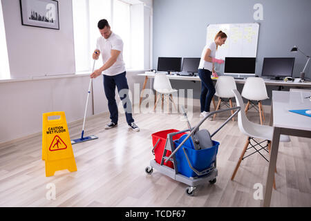 Two Smiling Young Janitor Cleaning The Desk And Mopping Floor In The Office Stock Photo