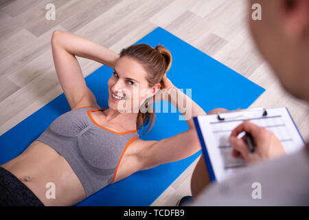 Sporty Young Woman Doing Sit Ups Over Yoga Mat Beside Trainer Holding Clipboard Stock Photo