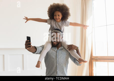 Happy african dad wearing crown piggybacking little daughter taking selfie Stock Photo