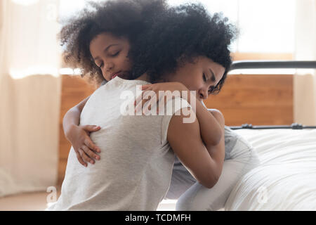 Cute small african american daughter hugging upset mother on bed Stock Photo