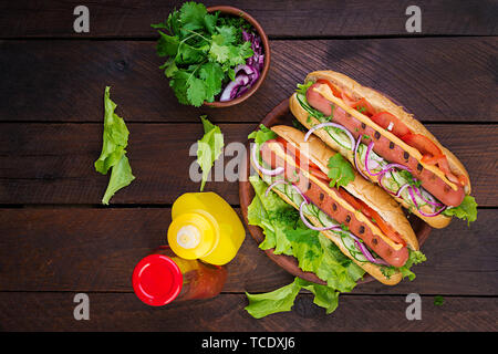 Hot dog with  sausage, cucumber, tomato and lettuce on dark wooden background. Summer hotdog. Top view Stock Photo