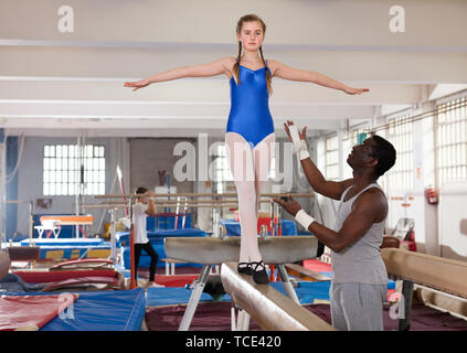 Happy African male coach helping teenage girl doing gymnastic exercises on equipment Stock Photo