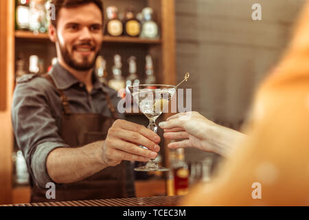 Handing a cocktail. Attractive smiling nice-looking dark-haired young-adult bar worker handing a martini cocktail to a female customer Stock Photo