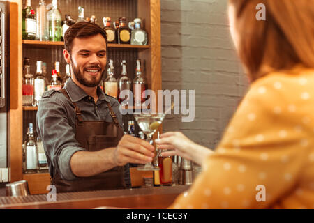 Giving a martini cocktail. Attractive beaming good-looking dark-haired adult bartender wearing dark apron giving a martini cocktail to a woman Stock Photo