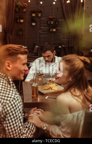 Flirting friends and lonely guy. Happy smiling cheerful good-looking young-adult radiant cheerful man and woman flirting with each other while their b Stock Photo