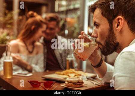 Feeling like the third wheel. Close-up face portrait of dark-haired bearded handsome young-adult man lonely sipping a cocktail while his mates hugging Stock Photo