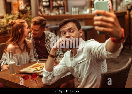 Taking selfies. Upset bored sad pessimistic sorrowful blue dark-haired nice-looking bearded man in his 25s taking selfies while his friends hugging at Stock Photo