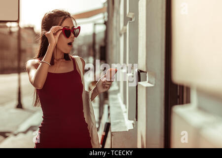 Shocked woman. Dark-haired slim hot nice-appealing bewitching woman in trendy eyeglasses and red dress feeling shocked while walking the streets Stock Photo