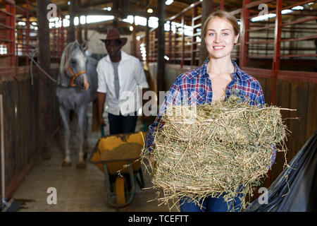 Two farm workers afro man and European girl spreading hay at stable Stock Photo