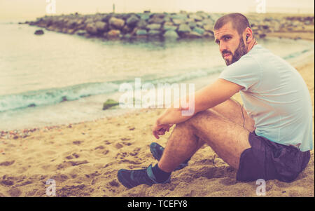 Young tired man resting, sitting on the sand near the sea after training Stock Photo