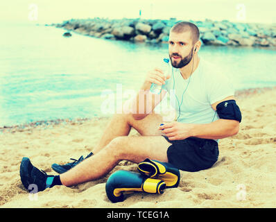 Young tired man resting, sitting on the sand near the sea after training Stock Photo