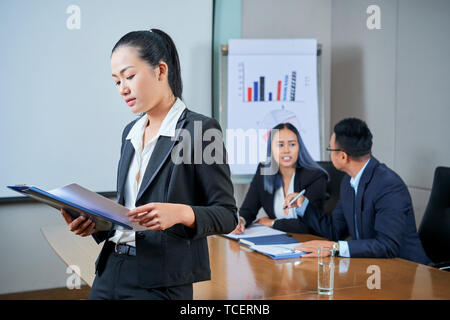 Young Asian businesswoman reading documents and getting ready for presentation when her colleagues taking at meeting table in background Stock Photo