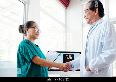 Smiling aged cardiologist shaking hand of female surgeon in green scrubs Stock Photo