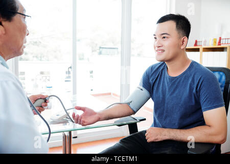 Doctor measuring blood pressure of young smiling Asian man Stock Photo